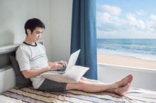 Man Works From Home Using Laptop In Bedroom With Beach View Through The Window