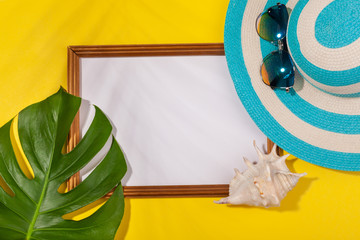 Wall Mural - Top view on striped blue hat, sunglasses, monstera leaf, seashell and frame for vacation photos on yellow background. Concept of beach holiday, sea tour, warm sunny summer. Copy-paste