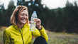 Happy sport girl laughing quenches thirst after fitness. Smile person drinking water from plastic bottles after exercising sport outdoors, woman isolation training in forest on nature, healthy