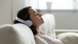 © fizkes - Peaceful girl in modern wireless headphones sit relax on comfortable couch listening to music, happy calm young woman in earphones rest on cozy sofa, enjoy good quality sound, stress free concept