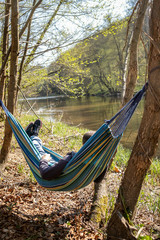 Young man is lying in a colorful hammock  near the trees, a river and mountainse early in the spring. He is alone and have a relaxing time in the nature