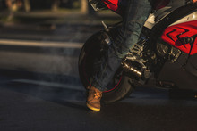 Crop View Of Bikers Foot And Back Wheel Of Red Sports Motorocycle. The Wheel Turns There Is Smoke From Burnt Rubber
