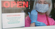 With cautious optimism a mature small business owner wearing a face mask and disposable gloves posts safety rules as she reopens her store after the coronavirus shutdown.