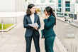 © Mangostar - Professional businesswomen discussing papers. Full length view of female colleagues in formal wear standing on street and discussing work. Paperwork concept