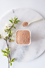 Ceramic bowl with red clay powder, for homemade facial and body mask or scrub and fresh sprig of flowering cherry on white background. Spa and bodycare concept.