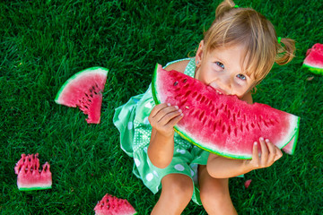 Little blonde girl sitting on the grass and eating slice of watermelon in summer.