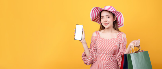 Beautiful asian woman in dress are holding shopping bags and mobile phone with face happily in yellow seamless background. Summer hopping lifestyle, online shopping, financial technology concept.