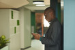 © AnnaStills - Young African businessman in suit standing at office corridor and typing a message on mobile phone