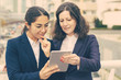 © Mangostar - Smiling businesswomen using tablet pc. Cheerful focused female colleagues standing with digital tablet on urban city street. Wireless technology concept