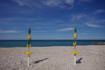 Two yellow-green umbrellas closed on a pebble beach, in the background the sea and the blue sky