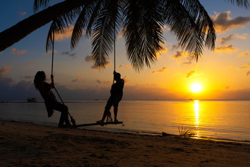 Silhouetted couple in love walks on the beach during sunset. Riding on a swing tied to a palm tree and watching the sun go down into the ocean