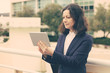 © Mangostar - Smiling businesswoman using tablet computer. Content middle aged businesswoman using digital tablet while standing on urban city street. Wireless technology concept