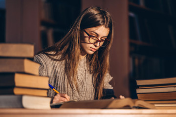 Young student in glasses preparing for the exam. Girl in the evening sits at a table in the library with a pile of books