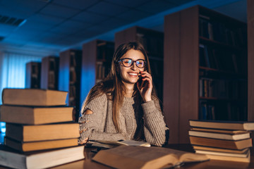 Young happy smiling student in glasses preparing for the exam. Girl in the evening sits at a table in the library with a pile of books, smiling and looking at the camera