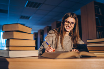 Young student in glasses preparing for the exam. Girl in the evening sits at a table in the library with a pile of books, smiling and looking at the camera
