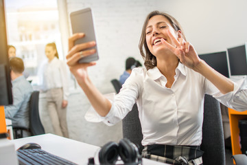 Wall Mural - Beautiful young woman taking selfie with smartphone and showing two fingers in office. Portrait of excited cheerful pretty woman showing v-sign or peace symbol with two fingers to mobile phone.