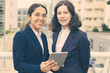© Mangostar - Content businesswomen with digital tablet. Cheerful professional female colleagues standing with digital tablet and smiling at camera at street. Wireless technology concept