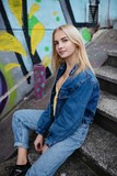 Fototapeta  - Young blonde woman smiling while sitting on ground against brick wall with graffiti.