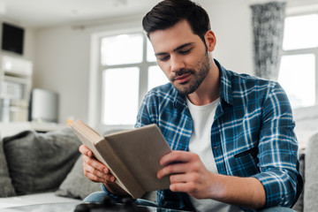 Selective focus of handsome man in plaited shirt reading book at home