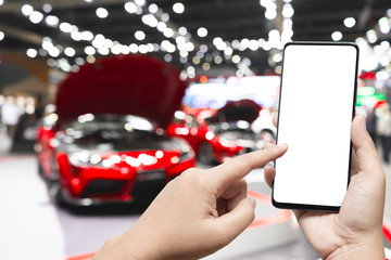 Mockup image of hand holding blank screen mobile and pointing to smart phone with blurred background of new cars display in showroom, buying new car. Mock up smartphone for your advertisement.