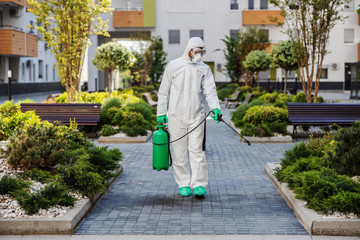 Wall Mural - Full length of man in sterile uniform and mask sterilizing surfaces in park from corona virus/ covid 19.