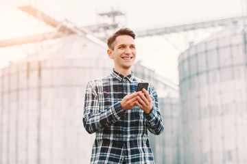 Portrait of happy young agriculture business owner holding mobile phone and smiling while standing against silos storage building. Successful confident business man owner of grain warehouse facilities