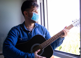 Bohemian musician with mask on his face and hat playing the Spanish guitar from the sofa. Dressed in a Hawaiian shirt and jeans. Concept music from home during confinement. Coronavirus pandemic.