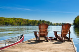 Fototapeta  - Two Adirondack chairs on a wooden dock on a lake in Muskoka, Ontario Canada. A red canoe is tied to the pier. Across the water cottages nestled between green trees are visible.