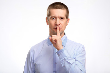 Wall Mural - successful businessman put his index finger to his lips, calling for a secret, showing a sign of silence, in a blue light casual shirt, a glance at the camera. Isolated