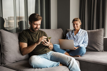 Wall Mural - Photo of joyful couple using laptop and mobile phone while sitting