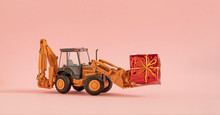 Toy Excavator, Backhoe Wheel Loader, Carries A Huge Gift Tied With A Bow. Pink Background. Copy Space. 