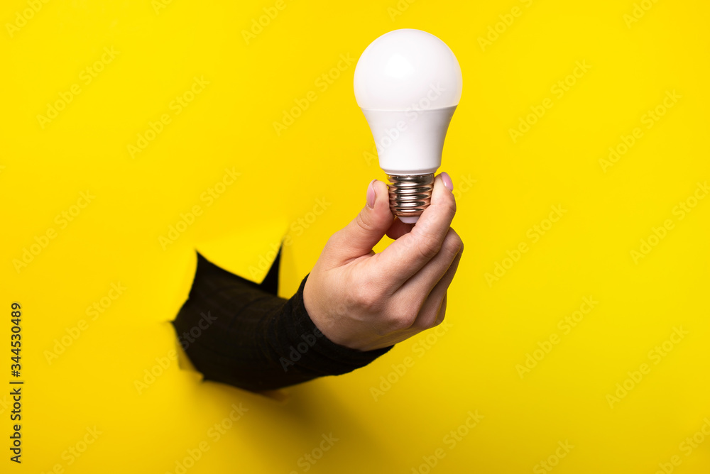 Hand holding an incandescent led light bulb from a torn hole in yellow paper - obrazy, fototapety, plakaty 