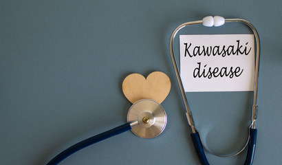 White card with words 'kawasaki disease' and stethoscope on blue background. Wooden heart.