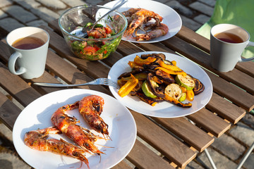 grilled langoustines and vegetables on white plates, a bowl with fresh salad, mugs of tea on a wooden table outside. Healthy lunch for two. Summer time