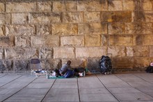 Homeless Person Sitting On Sidewalk Against Stone Wall