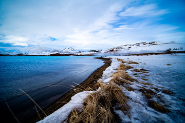 A thin strip of black beach in Iceland reaching into the horizon next to a turquoise blue lake. A large pointy mountain in the distance. Geothermal smoke blowing. Blue but cloudy sky.