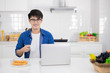 © Jedsada - Asian freelance young man working from home with breakfast bread and coffee using laptop in white kitchen