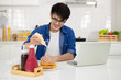 © Jedsada - Asian freelance young man working from home with breakfast bread and coffee using laptop in white kitchen