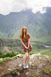 young beautiful girl with red hair in a red dress stands on a background of mountains in Georgia