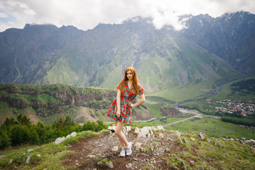 young beautiful girl with red hair in a red dress stands on a background of mountains in Georgia