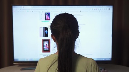 Back view of brunette female, which looks online shop on a big display PC and selects a mobile phone  to order online.