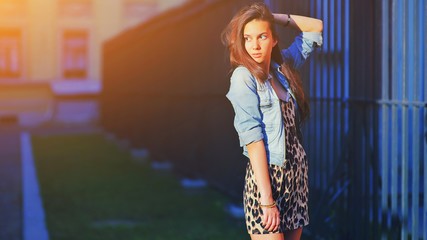 Stylish long hair brunette woman in a leopard animal print dress in blue denim jacket, street fashion concept. Fashionable hipster girl posing on summer evening street. Trendy boho style outfit.