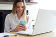 © grc - Smiling young female entrepreneur reading emails on her cellphone while working on a laptop at home. Telecommuting lifestyle.