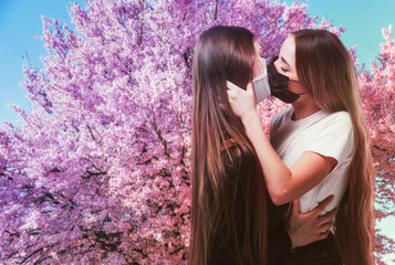 Lesbians in medical masks hug and kiss against the background of flowering trees