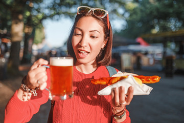 Cheerful Asian girl enjoys snacking on junk food - hot dog with fried sausage and beer