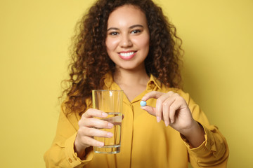 Wall Mural - African-American woman with glass of water and vitamin pill against yellow background, focus on hands