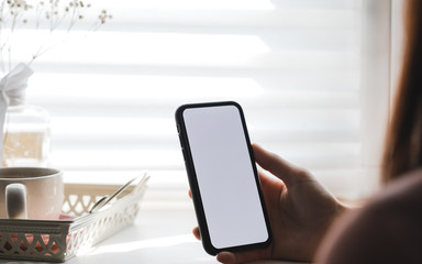 Woman uses a smartphone with a white screen, near the window at home in the morning. Mock up