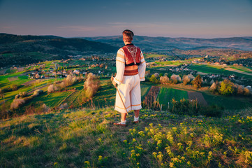 Wall Mural - A young man in a Slovak folk costume looks at the spring landscape in the village of Hrinova in Slovakia. Rising sun and spring flowering trees in the background. Edit Space.