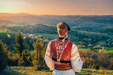 Wall Mural - Young man in folk costume standing in spring nature at sunrise. Edit space.