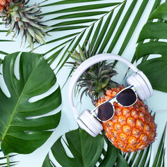 Wall Mural - Funny pineapple wearing white headphone, concept of listening music, isolated on blue background with tropical palm leaves, top view, flat lay design.